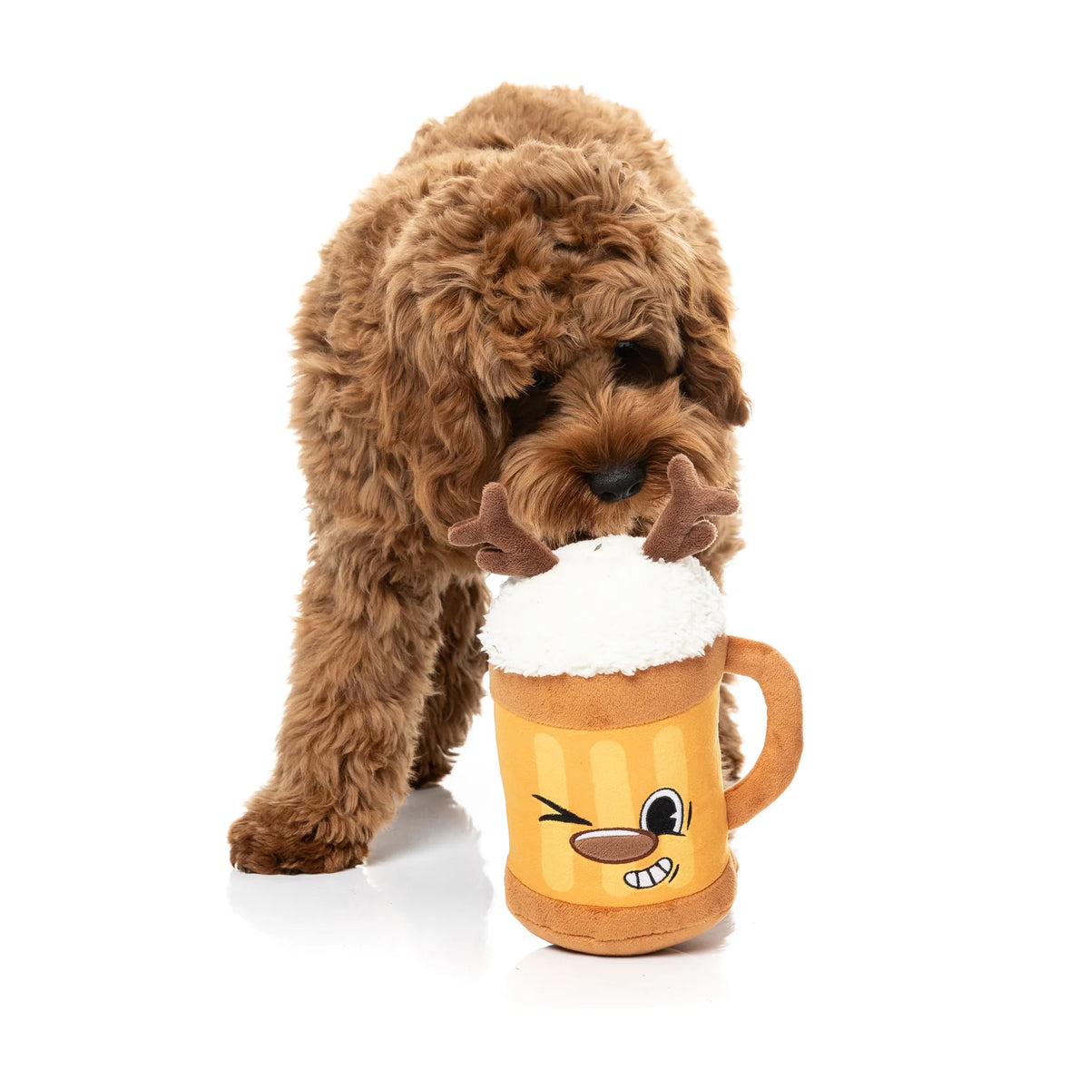 A brown curly-haired dog is holding a plush FuzzYard Jolly Reinbeer reindeer Christmas dog toy with antlers and a white faux-froam top, photographed on a clean white studio background.