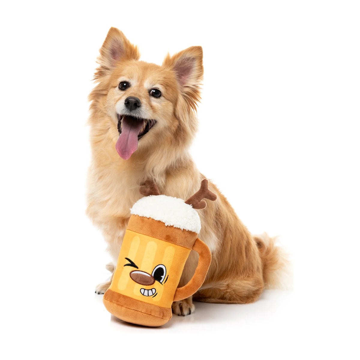 A small fluffy tan dog sitting on a white studio background with its tongue out, holding a plush FuzzYard Jolly Reinbeer reindeer Christmas dog toy with a cartoon face and small antler details.