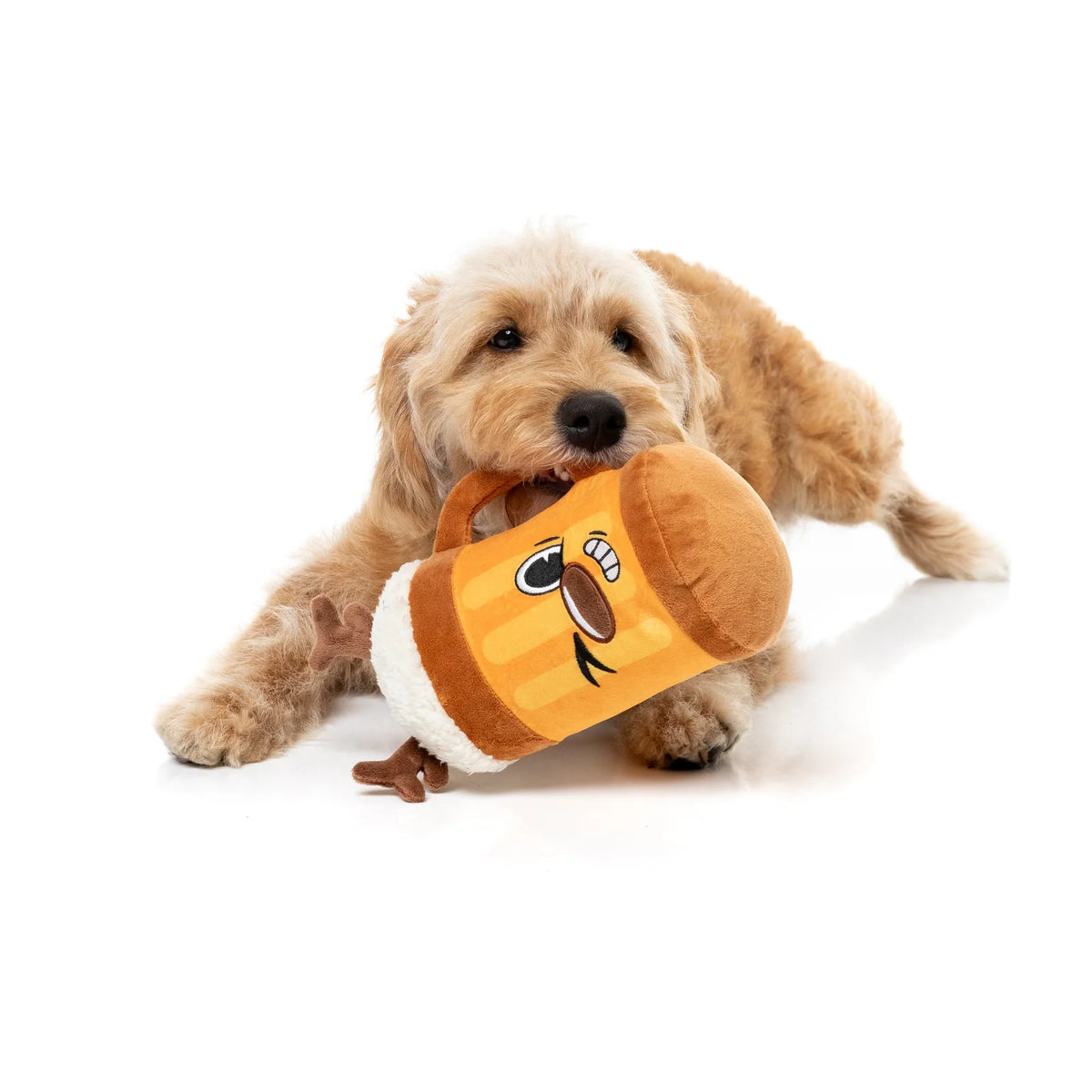 A small fluffy golden dog lying on a white studio background and biting a plush FuzzYard Jolly Reinbeer reindeer Christmas dog toy shaped like a beer mug with a cartoon face.