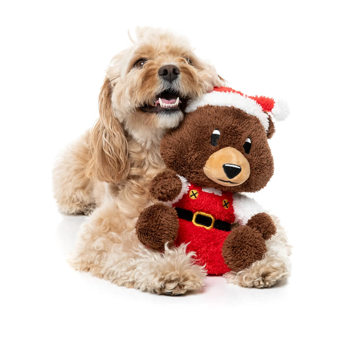 A fluffy cream-colored dog sitting behind a brown FuzzYard Santa Fuzz Bear plush Christmas dog toy wearing a red Santa hat and Santa suit, photographed on a clean white background.