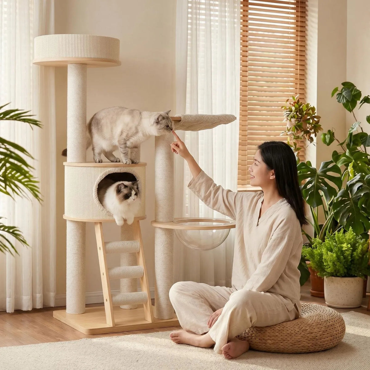 A woman in a beige outfit sits on a woven floor cushion next to a tall Petoria Grove Ladder cat tree with two cats, one on a platform and one in a cave, in a bright, plant-filled room.