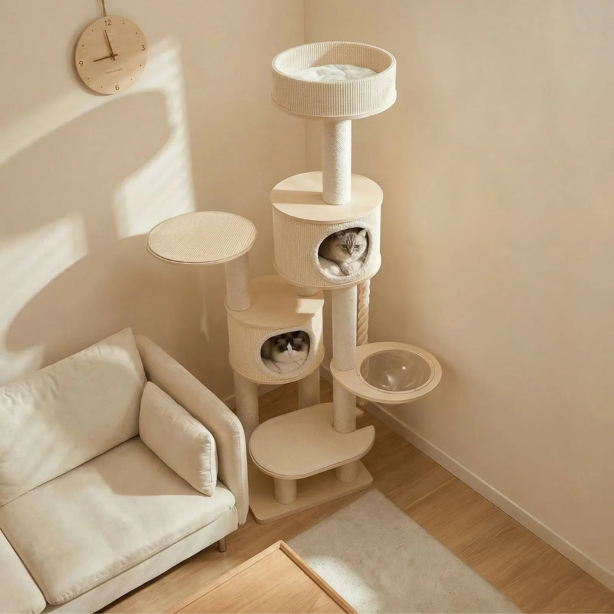 A tall cream-colored Petoria Grove Twin-Den Cat Tree with multiple levels, including a top perch, scratching posts, two enclosed cat beds with cats peeking out, and a clear bowl, placed in a sunlit living room next to a matching cream sofa.