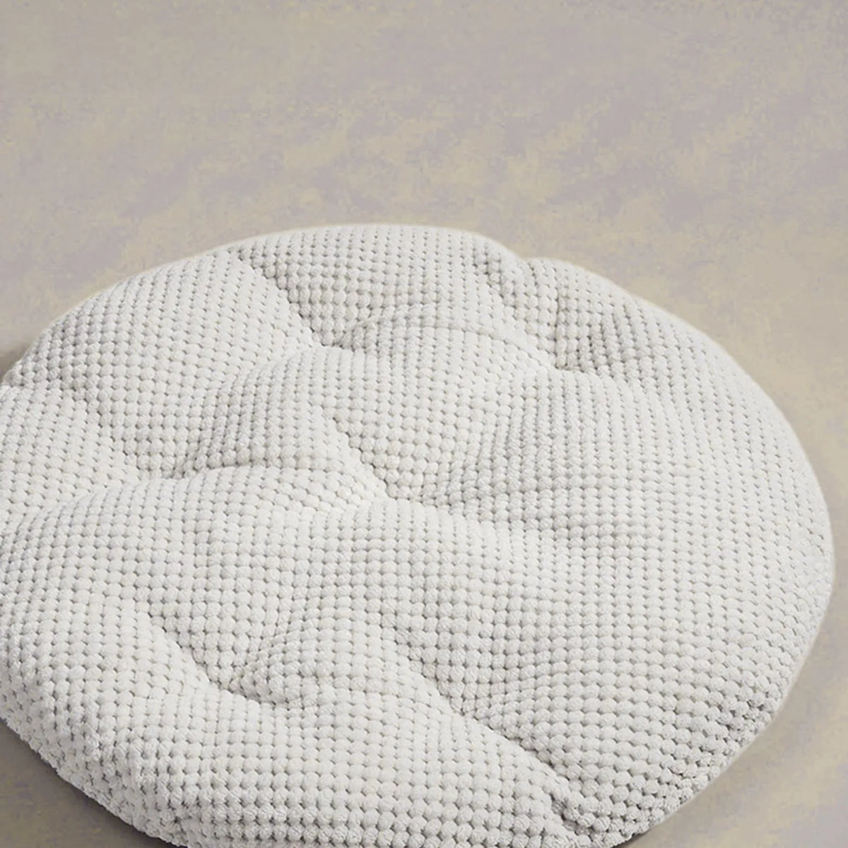 A round, off-white textured pouf or cushion with a waffle-weave pattern, photographed on a neutral beige background.