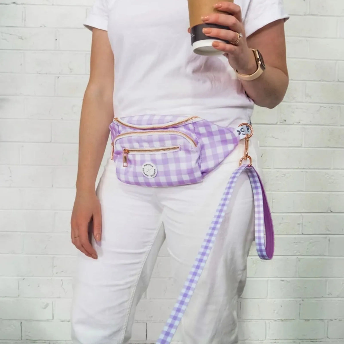 A person wearing a white t-shirt and white pants models a Pupstyle Berry Milkshake Dog Parent Bum Bag with a matching leash strap. The fanny pack has a front zippered pocket and a small round white logo patch, set against a white brick wall background.