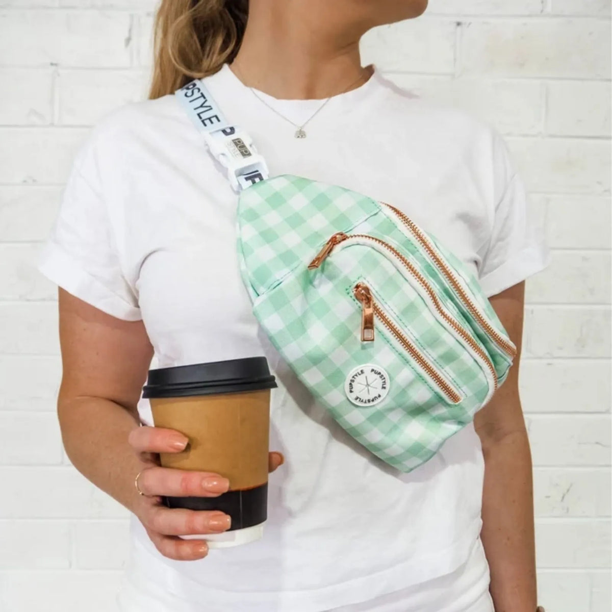 A woman wearing a Pupstyle Sweet Apple Dog Parent Bum Bag with adjustable waist strap crossbody with copper-toned zippers, holding a paper coffee cup against a white brick wall background.
