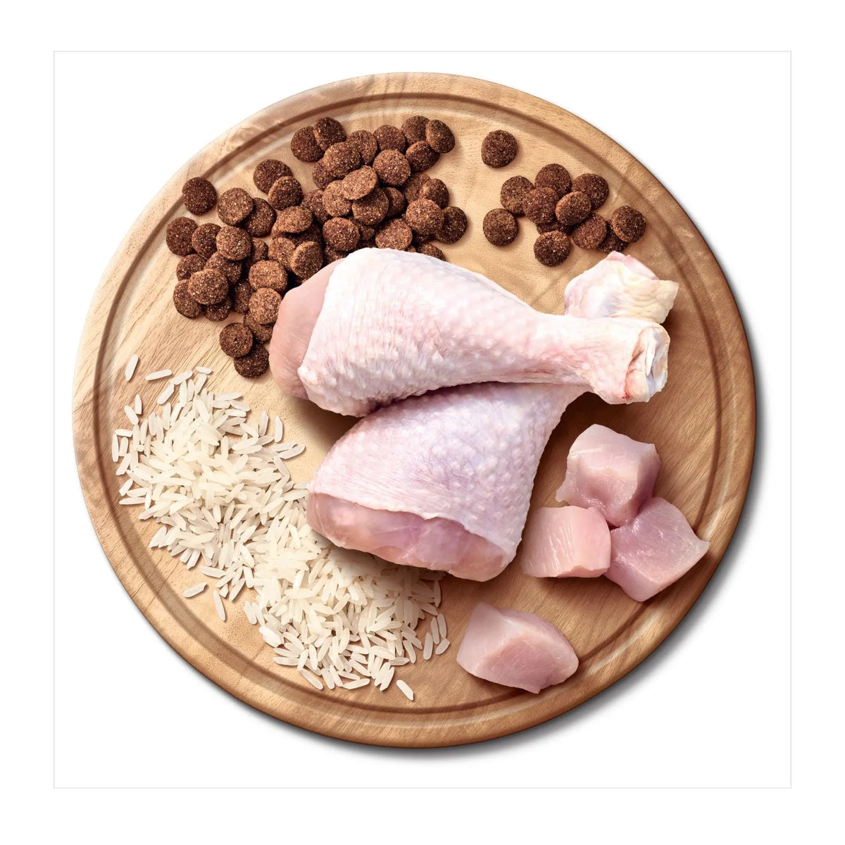 Raw chicken pieces, rice grains, and brown kibble dog food arranged on a round wooden cutting board against a white background.