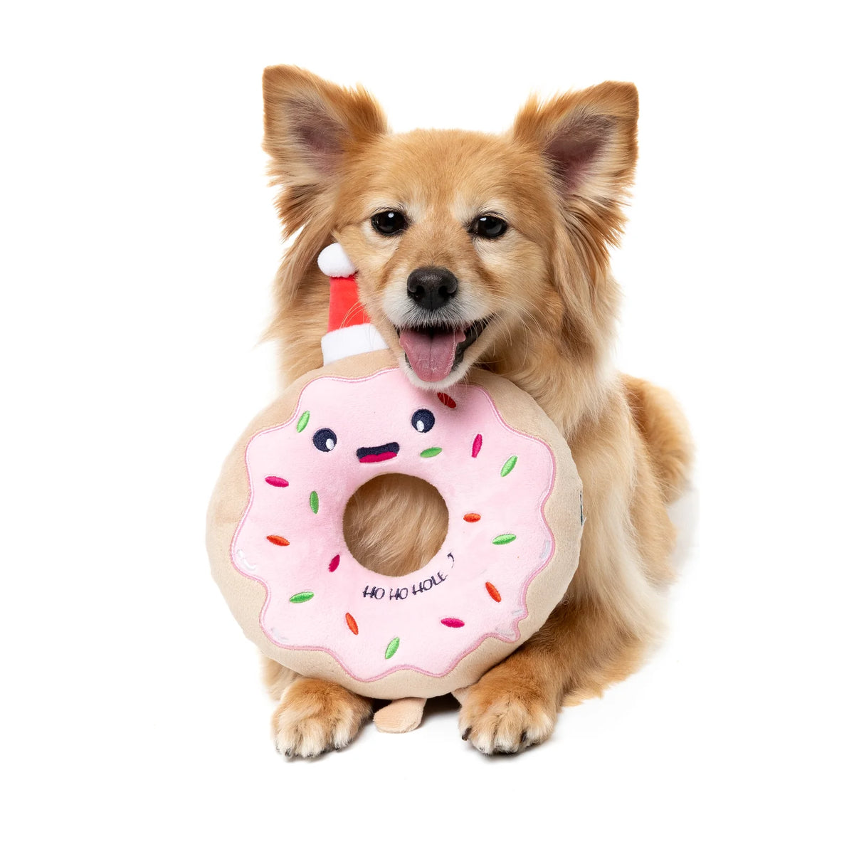 A small tan dog sits on a white background holding a plush FuzzYard Ho Ho Donut Christmas Dog Toy with pink frosting, colorful sprinkles, a smiling face, and a Santa hat.