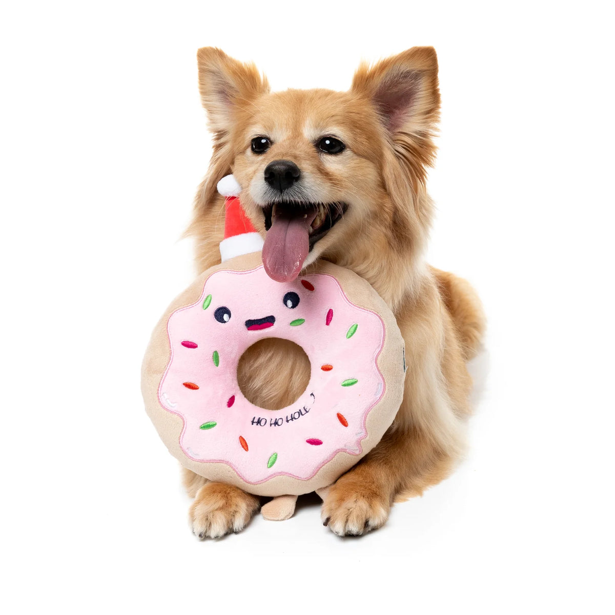 A small tan dog holding a plush FuzzYard Ho Ho Donut Christmas Dog Toy with pink frosting, colorful sprinkles, and a Santa hat, photographed on a clean white background.