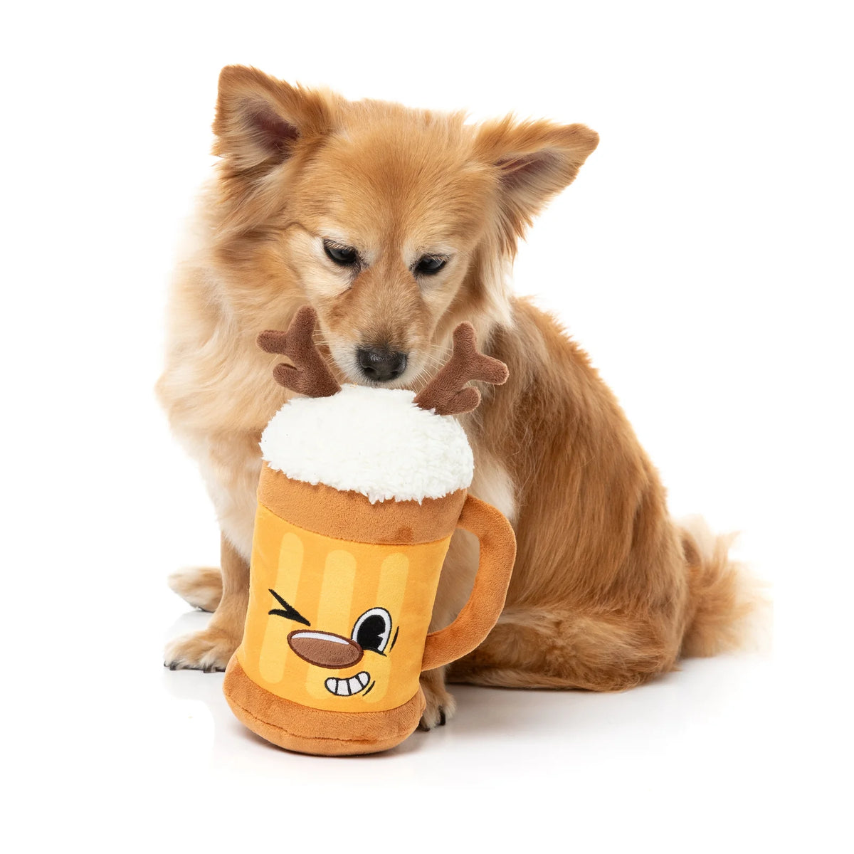 A fluffy golden-brown dog sits on a white studio background next to a plush FuzzYard Jolly Reinbeer reindeer Christmas dog toy with antlers and a white faux-foam top.