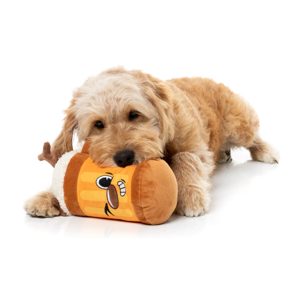 A small fluffy tan dog lying on a white studio floor and chewing on a plush FuzzYard Jolly Reinbeer reindeer Christmas dog toy with a cartoon face. The toy is orange and brown with white and black facial details.