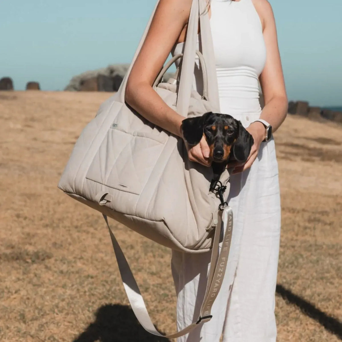 A woman in a white dress carries aFuzzYard Life Multipurpose Pet Carrier and Car Seat outdoors in a dry grassy field, with a dachshund tucked inside the carrier.