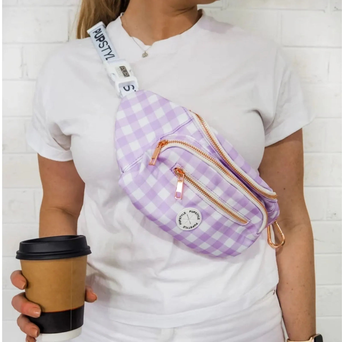 A person wearing a white t-shirt models a Pupstyle Berry Milkshake Dog Parent Bum Bag with rose gold zippers and a white adjustable strap. The person is holding a brown paper coffee cup against a white brick wall background.