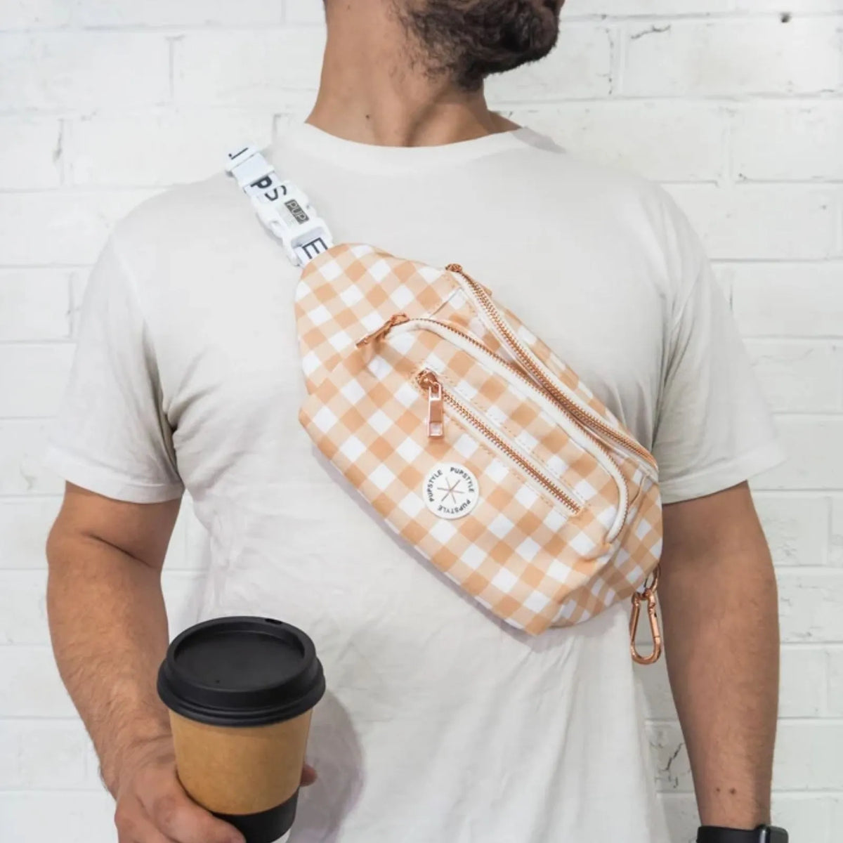 A man wearing a Pupstyle Creme Brulee Dog Parent Bum Bag  with a white strap and rose gold zipper, holding a coffee cup against a white brick wall.