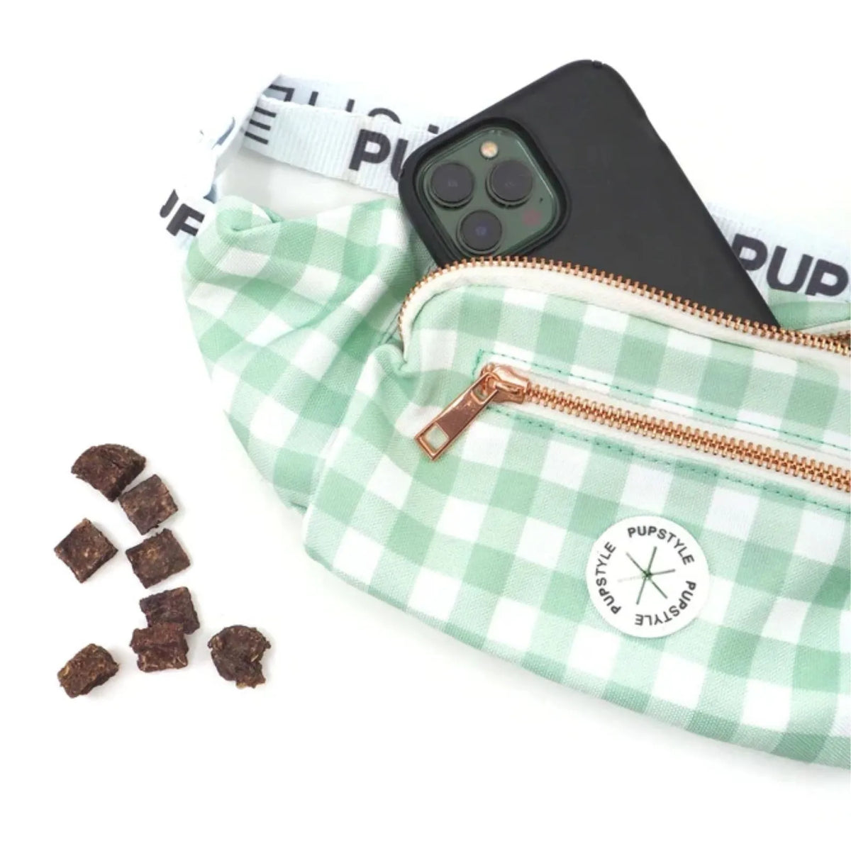 A Pupstyle Sweet Apple Dog Parent Bum Bag with a rose gold zipper, shown with a black smartphone tucked inside and small brown snack pieces on a white background. The brand name 'PUPSTYLE' is visible on the strap and the circular patch on the front.