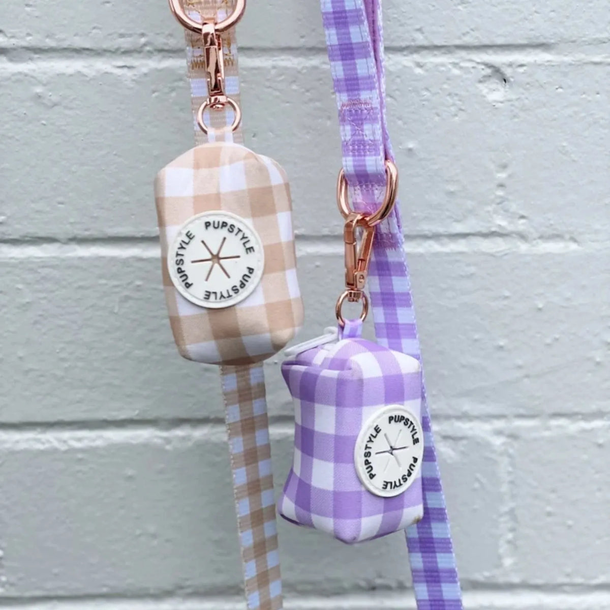 Two small fabric Pupstyle Berry Milkshake Dog Poop Bag Holder with matching leads hang against a light gray painted brick wall. 